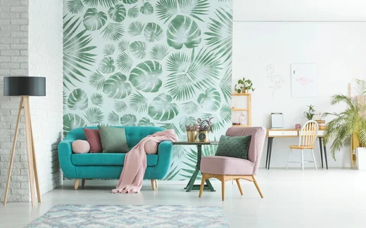Modernes Wohnzimmer mit grüner Blättertapete, türkisfarbenem Sofa, rosa Stuhl und Schreibtisch im Hintergrund.