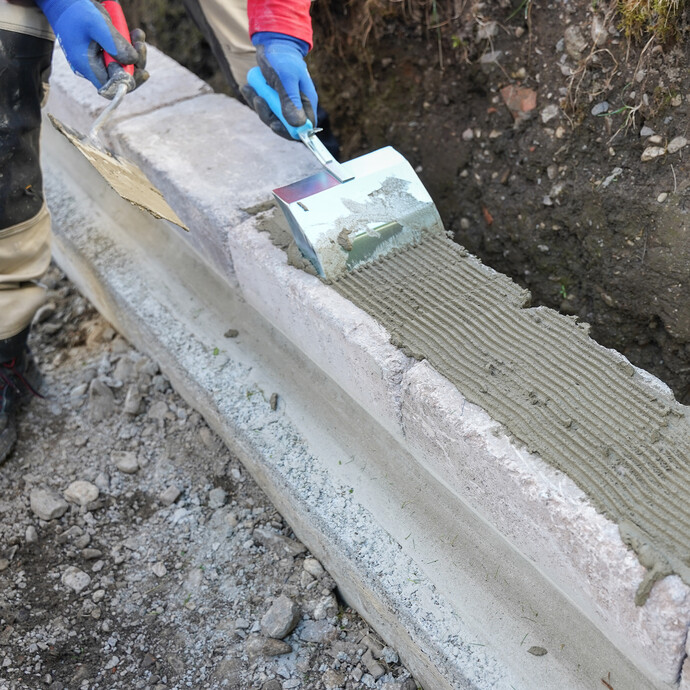 Eine Person kämmt frischen Steinkleber mit einer Klebekelle auf der Mauerfläche durch.