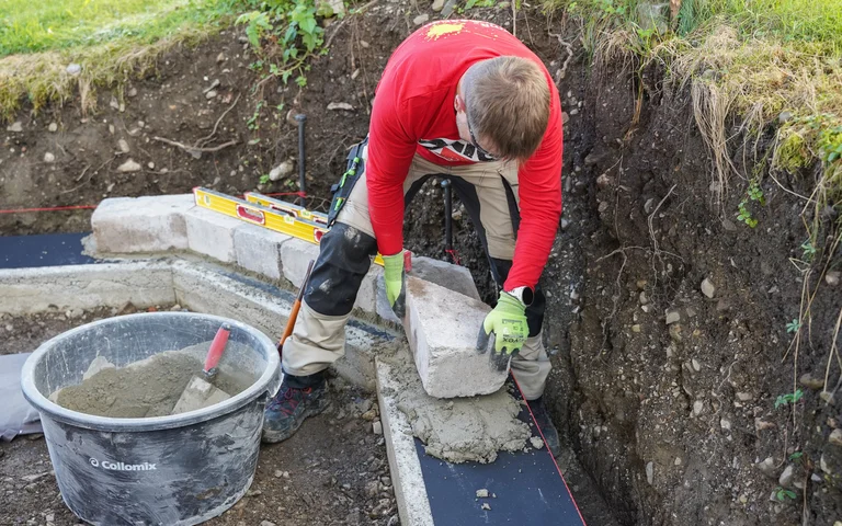 Eine Person setzt einen Mauerstein in einen frischen Mörtel auf das Fundament im Garten. Eine Wasserwaage und ein Mörtelkübel mit angerührtem Universalmörtel und Kelle liegen daneben.