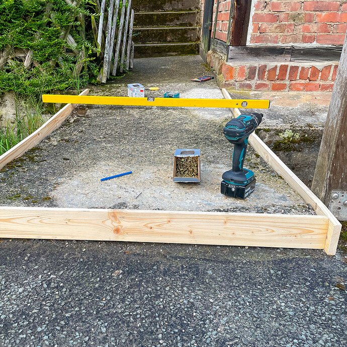 Holzschalung aus Kanthölzern wird auf dem Untergrund ausgerichtet und mit Wasserwaage kontrolliert, Akkuschrauber und Schrauben liegen bereit.