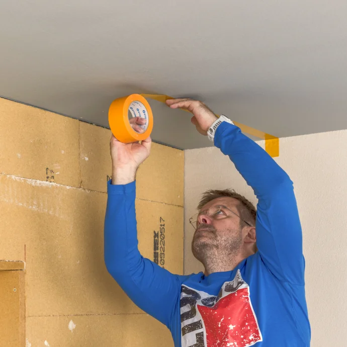 Heimwerker bringt Klebeband an einer Deckenkante an, um die Farben sauber zu trennen. Ideal für präzise Malerarbeiten.