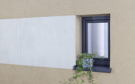 Fassade mit beigem Oberputz und verputzter Fläche in Betonoptik neben einem Fenster mit Blumenschmuck.